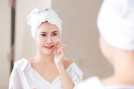 Young asia woman applying foundation or moisturizer on her face in front of mirror.の写真素材