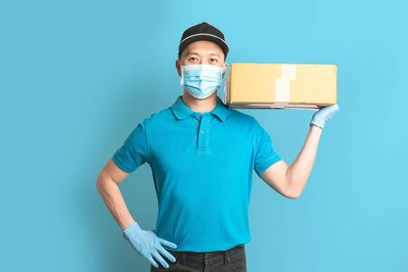 Delivery man employee in blue shirt uniform face mask gloves hold empty cardboard box on blue background. Coronavirus virus 2019 conceptの写真素材