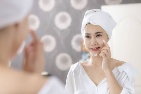 Young asian woman applying foundation or moisturizer on her face in front of mirror.の写真素材