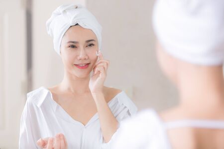 Young asian woman applying foundation or moisturizer on her face in front of mirror.