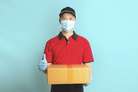 Delivery man employee in red shirt uniform face mask gloves hold empty cardboard box on blue background. Coronavirus virus 2019 conceptの写真素材