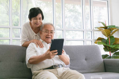 Couple of seniors smiling and looking at the same tablet hugged on the sofa.の写真素材