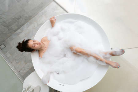 Asian beautiful young woman enjoying pleasant bath with foam, lying with closed eyes. Top viewの写真素材