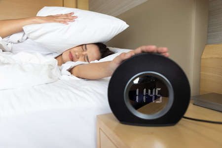 Sleepy young woman trying kill alarm clock while bury hand in pillow.の写真素材