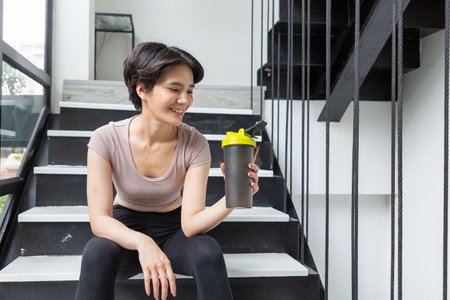 Athlete girll sitting in a black tight-fitting sports suit with a drinking bottle and sitting on the stairs.の写真素材