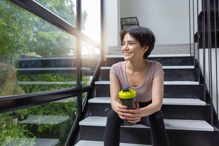 Athlete girll sitting in a black tight-fitting sports suit with a drinking bottle and sitting on the stairs.の写真素材