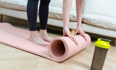 Close-up of attractive young woman folding pink yoga or fitness mat after working out at home in living room.の写真素材