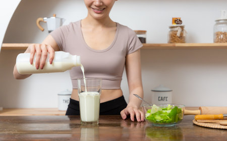 Smiling young woman pouring milk into a glass while sitting and having breakfast at the kitchen table.の写真素材