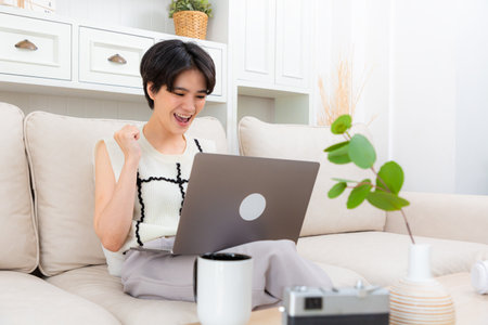 Smiling young woman using laptop, sitting on couch at home, beautiful girl shopping or chatting online in social network or freelancer working on computer project.の写真素材