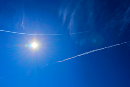 Bright sun with flares and spectral aureole against blue sky with white jet tracesの写真素材
