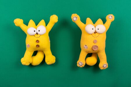 Two funny friendly smiling yellow cats with open paws against green background, designer soft toys