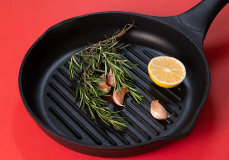 Black frying pan with rosemary sprigs and cloves of garlic inside against bright red backgroundの写真素材