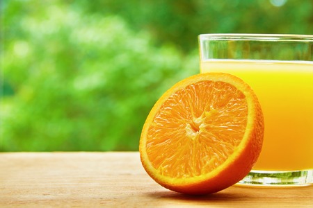 Photo of glass of orange juice and the half of orange on the wooden table on the green blurred backgroundの写真素材
