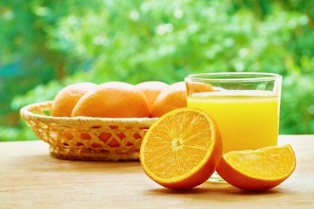 Glass of orange juice, half of orange, orange lobule and basket with oranges on the wooden table on the green blurred backgroundの写真素材