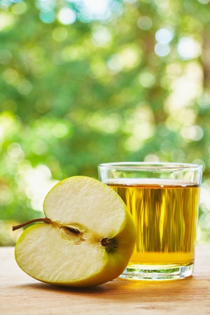 Glass with apple juice and halved apple on the wooden table on green blurred backgroundの写真素材