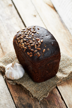 Close up photo of rustic bread sprinkled with sunflower seeds on sackcloth and garlic on planked...