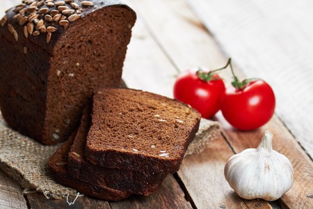 Close up photo of rustic bread sprinkled with sunflower seeds on sackcloth, garlic and two tomatoes on planked wooden tableの写真素材