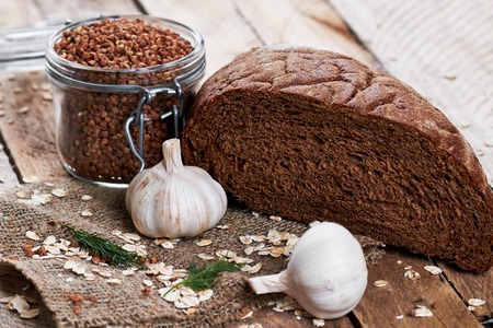 Close up photo of rustic bread on sackcloth, garlic, dill, oat flakes and jar with buckwheat on planked wooden tableの写真素材