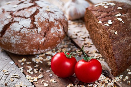 Closeup photo of two loaves of bread, garlic, two tomatoes and oat flakes on planked wooden tableの写真素材