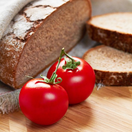Sliced loaf of gray floured bread, two tomatoes and two pieces of it on cotton towel on wooden cutting boardの写真素材