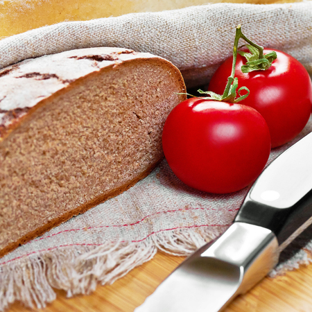Sliced loaf of gray floured bread, tomatoes and knife on cotton towel on wooden cutting boardの写真素材
