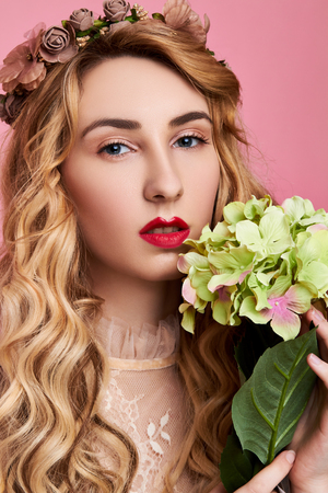 Fashion photo of young woman on pink background wearing flower wreath with flower near her faceの写真素材