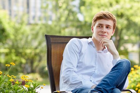 Young attractive man in white shirt sitting on brown chair in the park on green backgroundの写真素材