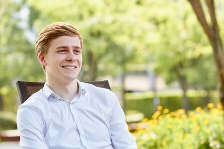 Young attractive man in white shirt sitting on brown chair in the park on green backgroundの写真素材