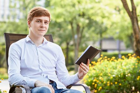Young attractive man in white shirt sitting on brown chair in the park on green backgroundの写真素材