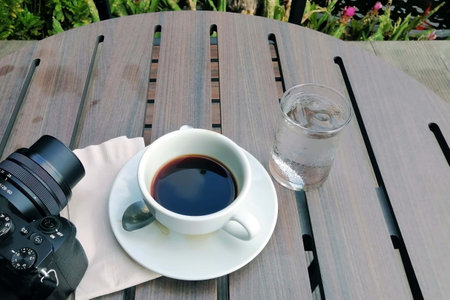 A black coffee cup on wooden table with camera and glass of water with a garden background.の写真素材