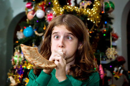 A young Christian girl blows a Jewish Shofar on Christmas morning.  It's a melding of religions and traditions as she stands in front of a Christmas tree.  She is blowing so far that her cheeks are puffed out and her eyes are slightly crossed.の写真素材