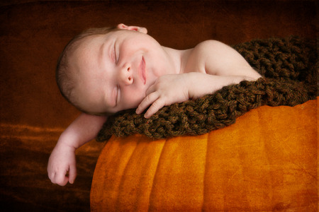 Happy newborn sleeping in pumpkin.  He is smiling in his sleep with his arm hanging over the edge of the pumpkin.  The image has been textured for a more artistic look.の写真素材