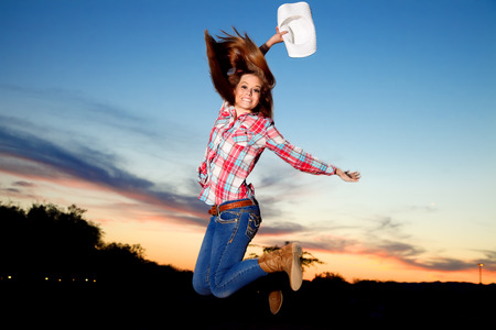 A teenage cowgirl jumps in the air at sunset.  She holds her western hat in her hand above her head in celebration.の写真素材