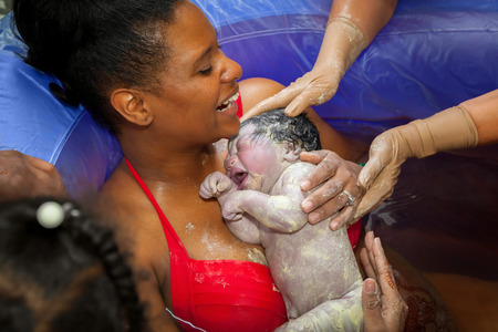 A happy African American mother holding her brand new baby girl after delivering her in a birthing pool at home.  The baby is covered in vernix and the mom is laughing.