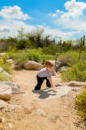 A cautious little boy hiking in the desert bends over to climb some low rocks on a trail.の写真素材