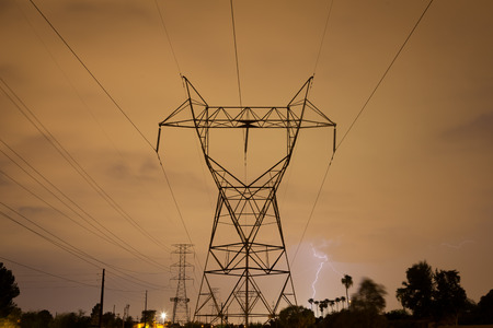 Lightning striking behind palm trees in the distance.  A power tower and lines stand tall in the foreground.  It is a stormy, summer night in the Arizona desert during monsoon season.の写真素材