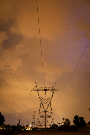 Lightning striking behind palm trees in the distance.  A power tower and lines stand tall in the foreground.  It is a stormy, summer night in the Arizona desert during monsoon season.の写真素材