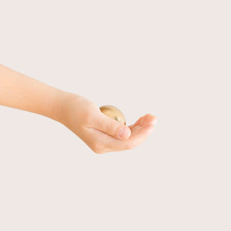 Child's hand holds gold Easter egg on the pastel beige background with copy space. Minimal creative, festive, luxury concept.の写真素材