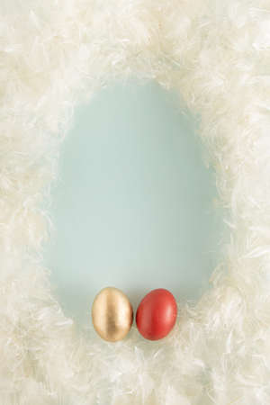 Soft fur pastel white and beige colors background made of fluffy seed hairs. Minimal style flat lay border frame with egg shaped copy space. Easter greeting card with red and gold Easter eggs.の写真素材