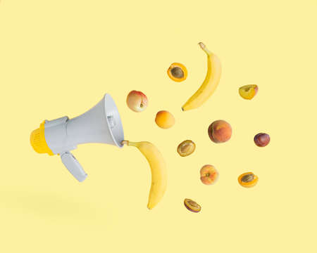 2021 Nature summer scene, made of gray megaphone and colorful summer fruits isolated on illuminating yellow background. Promotion raw, healthy food idea. Minimal, cute, aesthetic, advertising concept.の写真素材