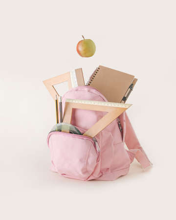 Pink school backpack full of books and supplies - triangles, rulers, pencils, notebook and apple fruit snack, isolated on pastel beige background. Minimal back to school idea.の写真素材
