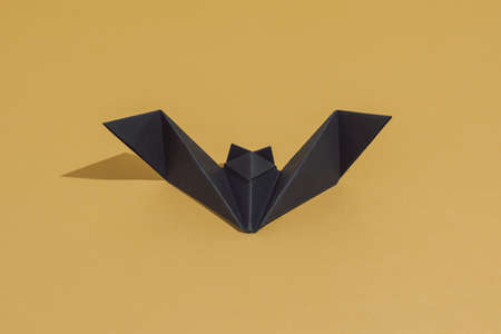 Origami bat made of black paper, isolated on gold or ocher background. Do it yourself decoration concept with copy space. Sun and shadows. Minimal luxury invitation card for Halloween party.の写真素材