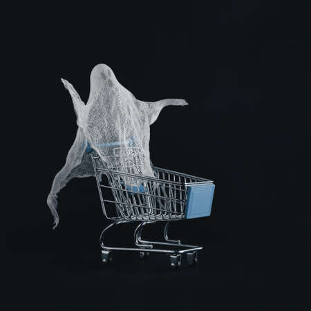 Minimal halloween scene made of pastel white ghost in a empty supermarket cart isolated on the black background. Halloween party purchase concept or consumer society ethos.の写真素材
