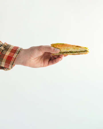 A male hand in a plaid earth-colored shirt holds slice of baklava cake with pistachios. Minimal delicious food scene isolated on pastel white background. Traditional oriental baked sweets concept.の写真素材