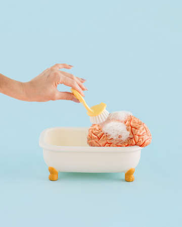 Young female hand brushing a human brain in a tub with foam, on isolated pastel blue background with copy space. Minimal abstract concept of mental health, treatment of brain fog or brainwashing.の写真素材