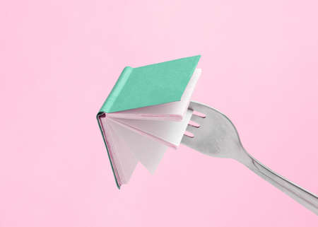 Minimal abstract composition made of fork holding a green book on isolated pastel pink background...