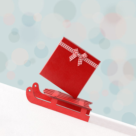 Red Santa's sleigh and gift box with ribbon and bow on snowy background with pastel bokeh. Minimal creative abstract Christmas card. New year poster. Holiday decoration concept.の写真素材