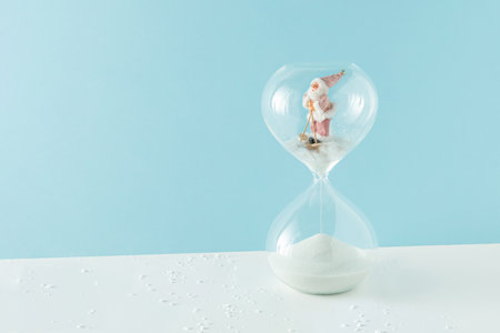 2023. Santa Claus on his skis in the snow in the hourglass on isolated pastel blue and white...