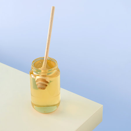 Jar of golden-yellow acacia honey with wooden honey dipper standing on beige table against isolated purple-blue background. Concept of natural, vegetarian, organic healthy food.の写真素材