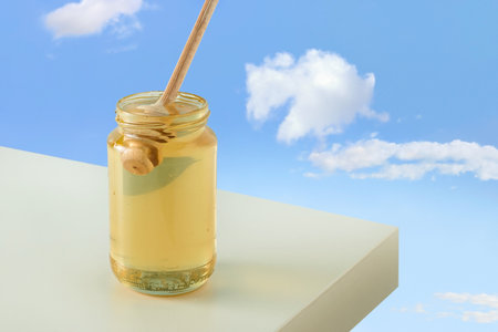 Glass jar with golden-yellow honey and wooden honey stick dipper on beige table. Isolated blue sky background. Concept of alternative medicine or traditional natural healthy food.の写真素材